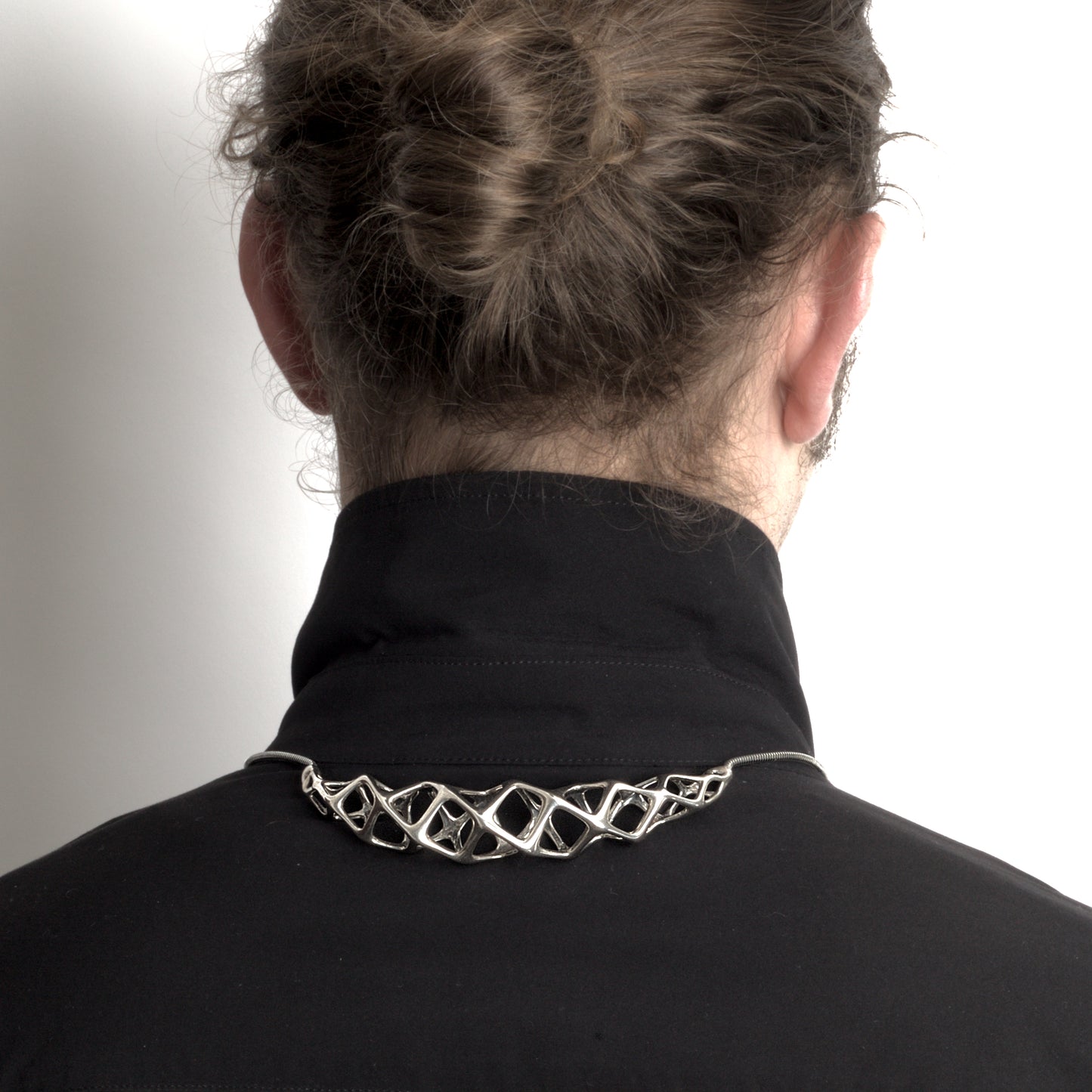 The solid sterling silver bolo tie–choker hybrid necklace styled on a man, with the necklace's geometric crest worn at the back of the shirt collar and the traditional bolo tie elements in front.