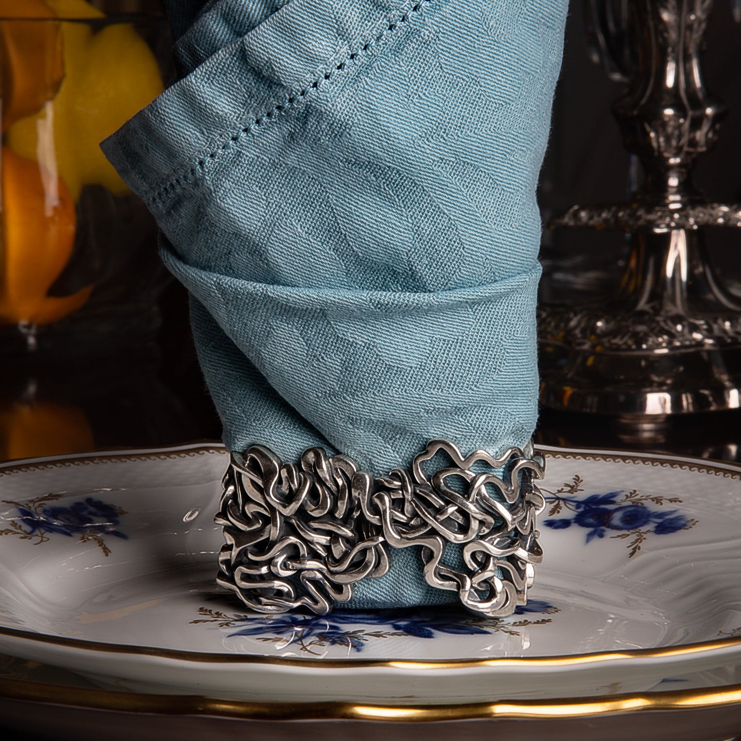 The rings can be styled in many ways using a variety of napkin folding techniques. In this image the ring is positioned upright creating the base for a flame folded napkin. This pairing is placed in the center of the dinner plates. Fresh picked lemons and a sterling candelabra are visible in the background and complete the scene.