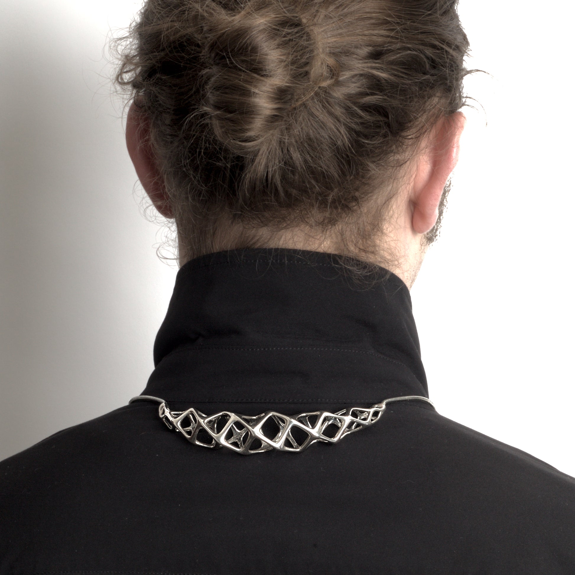 The solid sterling silver bolo tie–choker hybrid necklace styled on a man, with the necklace's geometric crest worn at the back of the shirt collar and the traditional bolo tie elements in front.