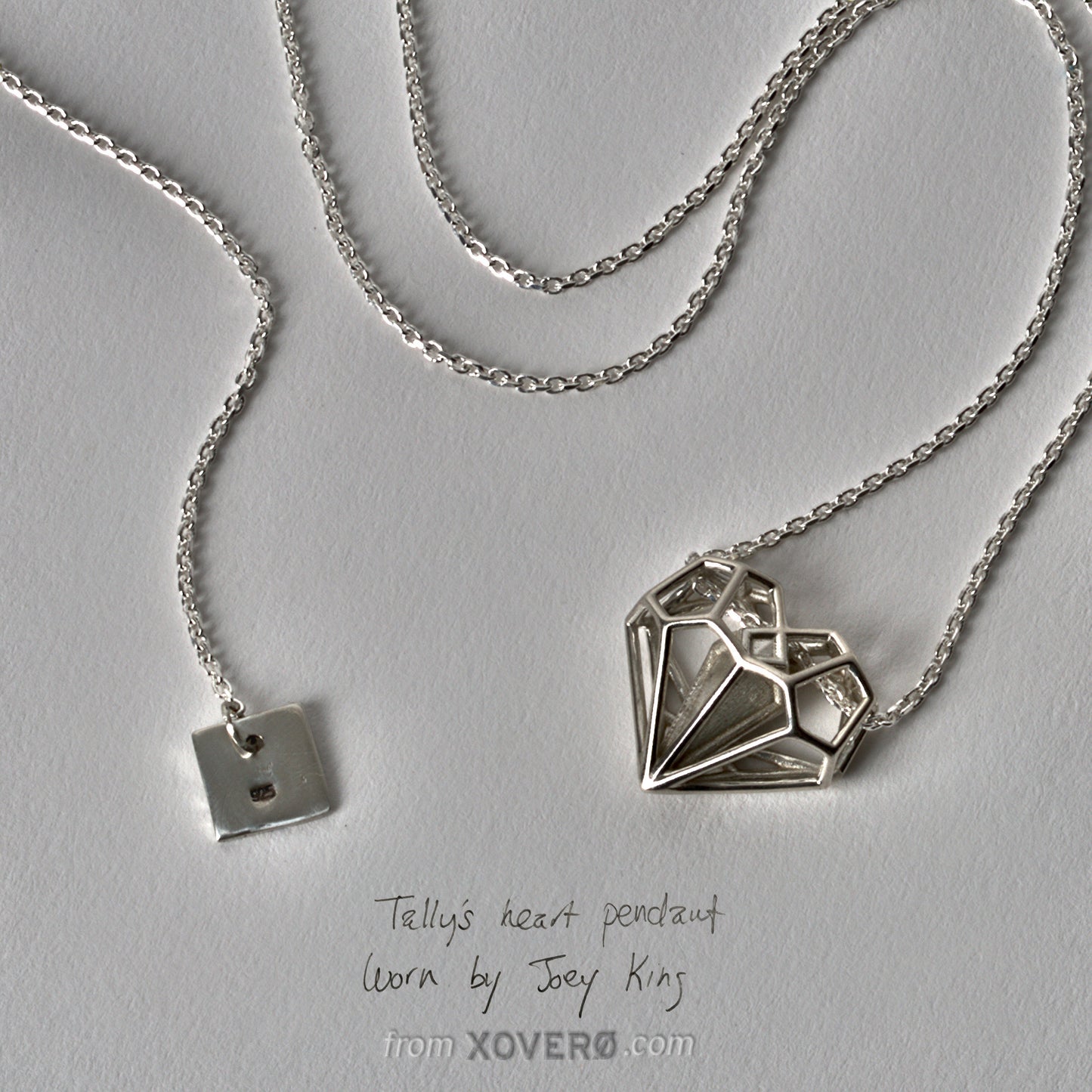 Detail of the Geoheart Pendant necklace from the Netflix movie Uglies, featuring a hallmark tag displaying the .925 sterling silver stamp. The pendant, designed by Xover0 Jewelry and worn by Joey King as Tally Youngblood, showcases a futuristic take on Art Deco architecture against a white paper background.