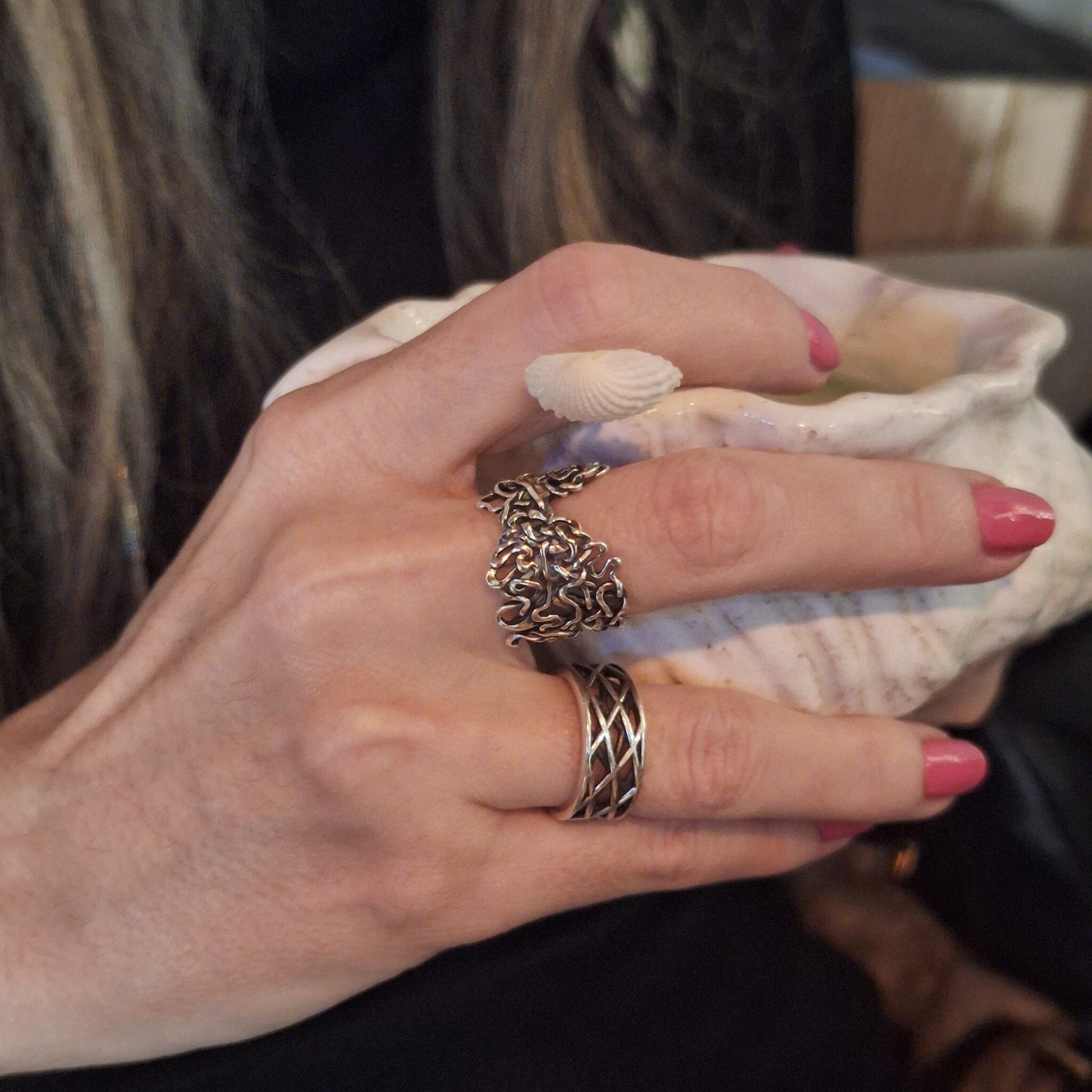 Hand wearing two silver rings with pink nail polish holding a shell. One ring features interlaced oxbows of the Mississippi River. 