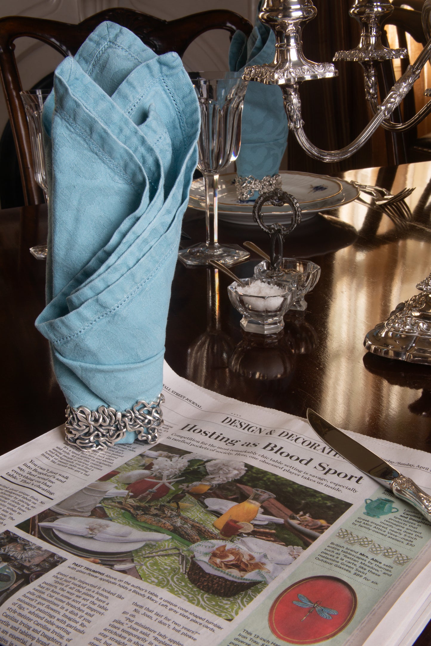 Xover0’s solid sterling silver Mississippi River napkin ring beside a Wall Street Journal article titled “Hosting as Blood Sport,” highlighting the competitive art of table setting.