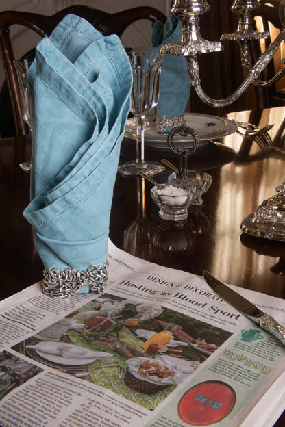 Xover0’s solid sterling silver Mississippi River napkin ring beside a Wall Street Journal article titled “Hosting as Blood Sport,” highlighting the competitive art of table setting.
